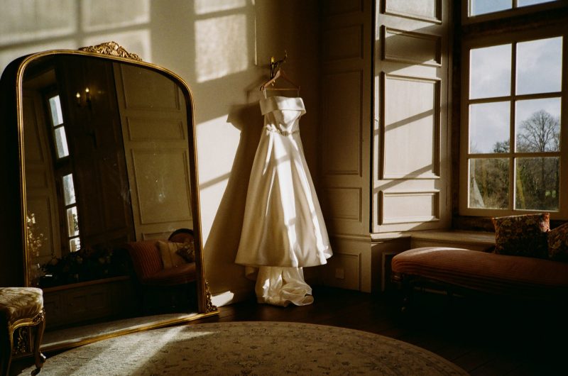 A wedding dress hangs beside a mirror in a room bathed in sunlight, near the wedding photobooth.