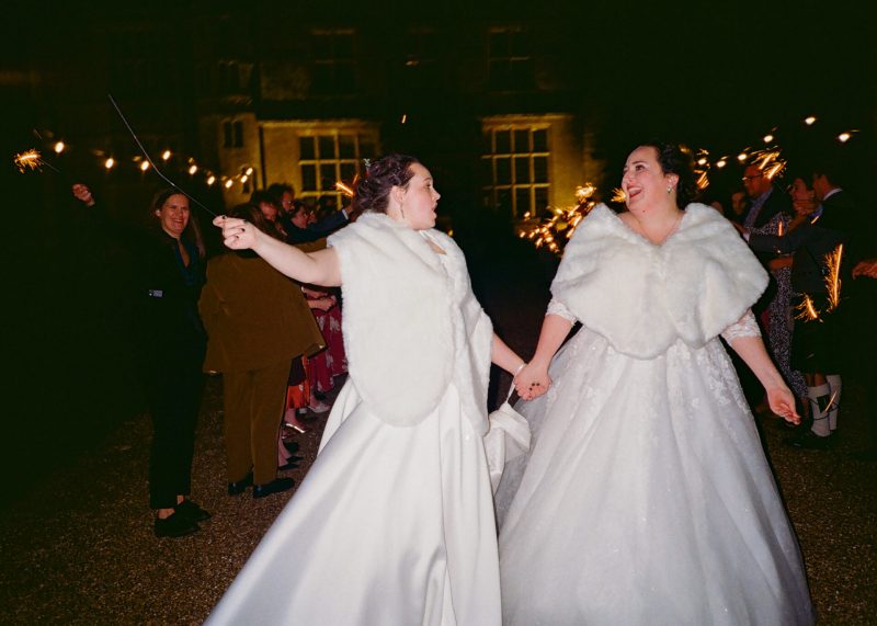 Two brides walk hand in hand through a sparkler send-off at their wedding at night.