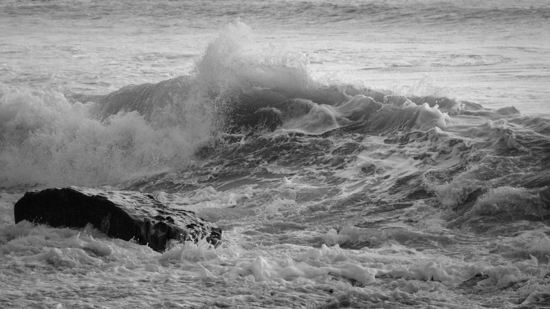 Waves crashing against a rock in a turbulent sea, captured in black and white.