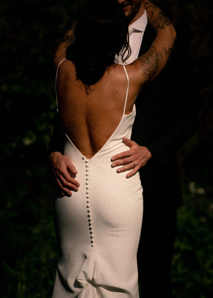 A couple in formal attire embracing, with focus on their hands and the back of the woman's dress.