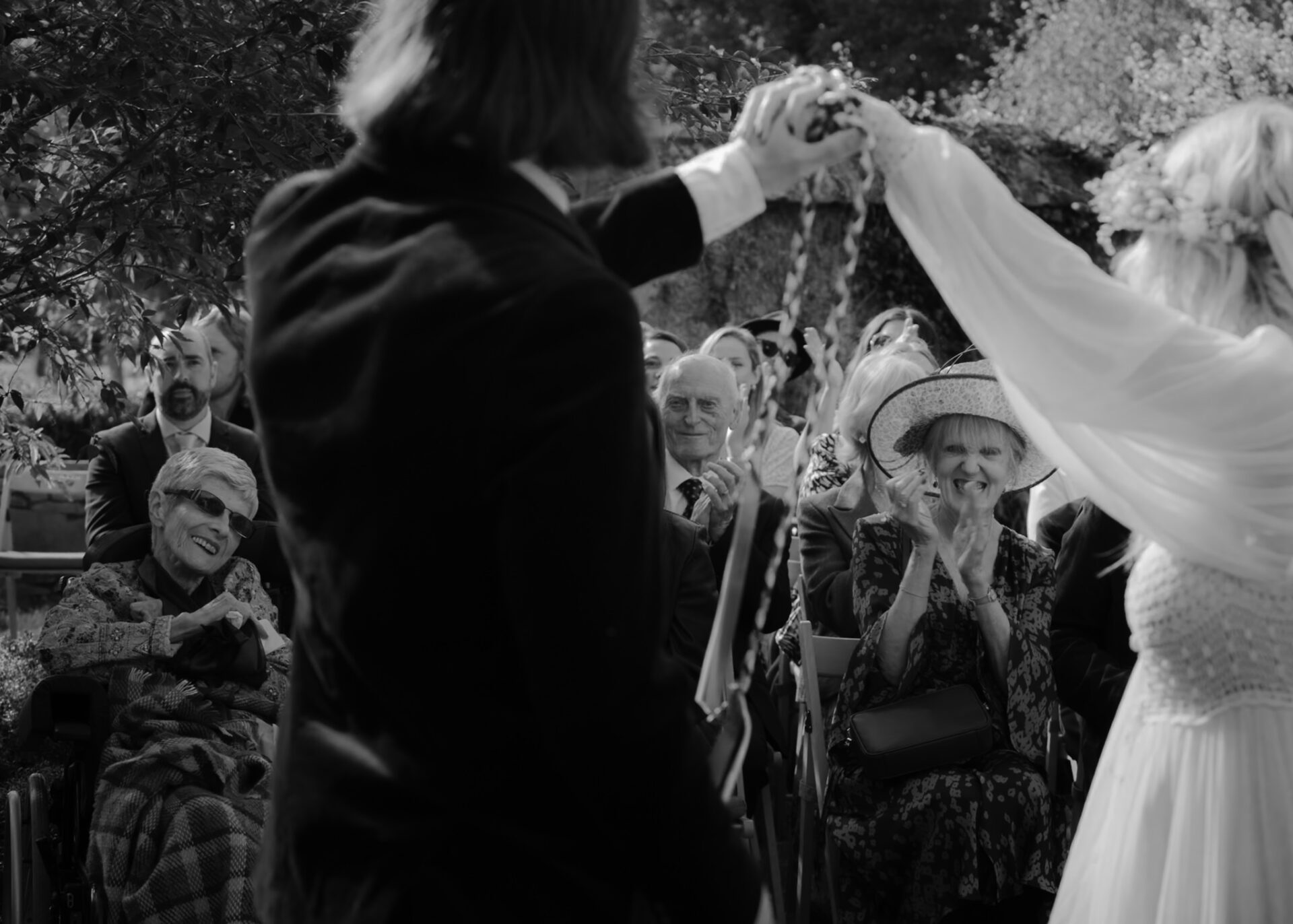 A black-and-white photo of a bride and groom holding hands at an outdoor wedding, with joyful guests watching, is part of our exclusive wedding photography collections.