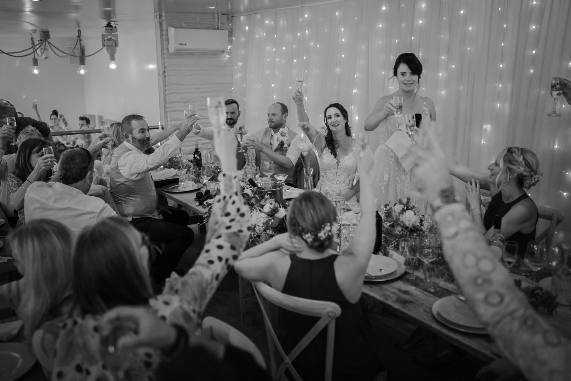 Guests at a wedding reception raising their glasses for a toast, with a joyful bride and groom at the center, surrounded by string lights and floral decorations during a photography session.