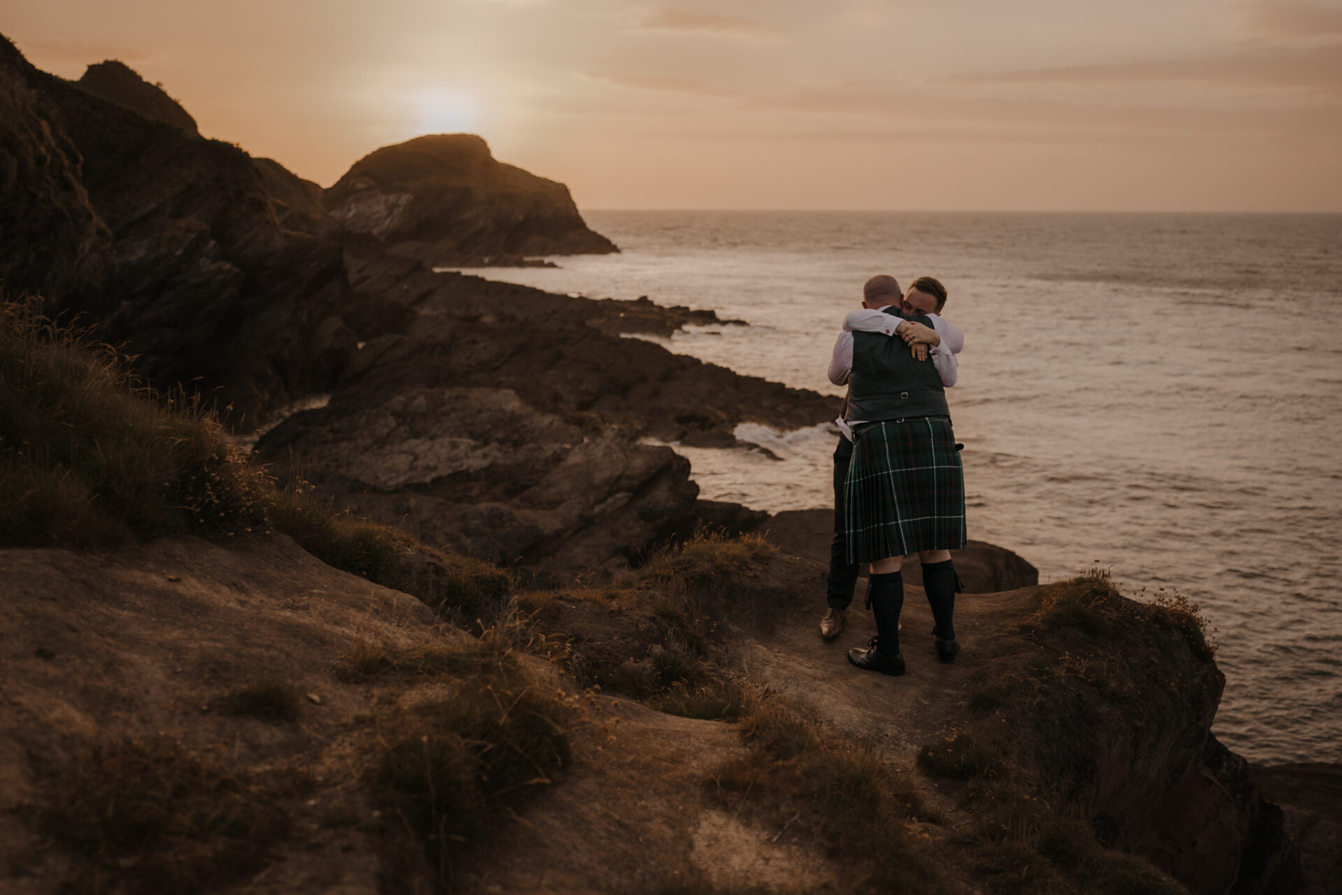Two people in kilts embracing by the rocky coast at sunset, with the ocean and cliffs in the background, reminiscent of a wedding collection.